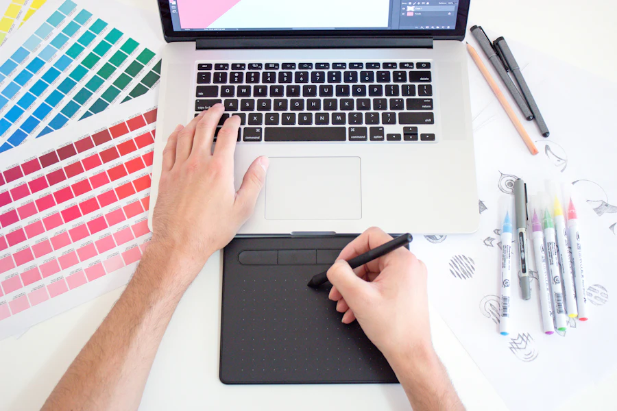 Designer working at a laptop and pen tablet beside fanned color swatches, markers and logo sketches