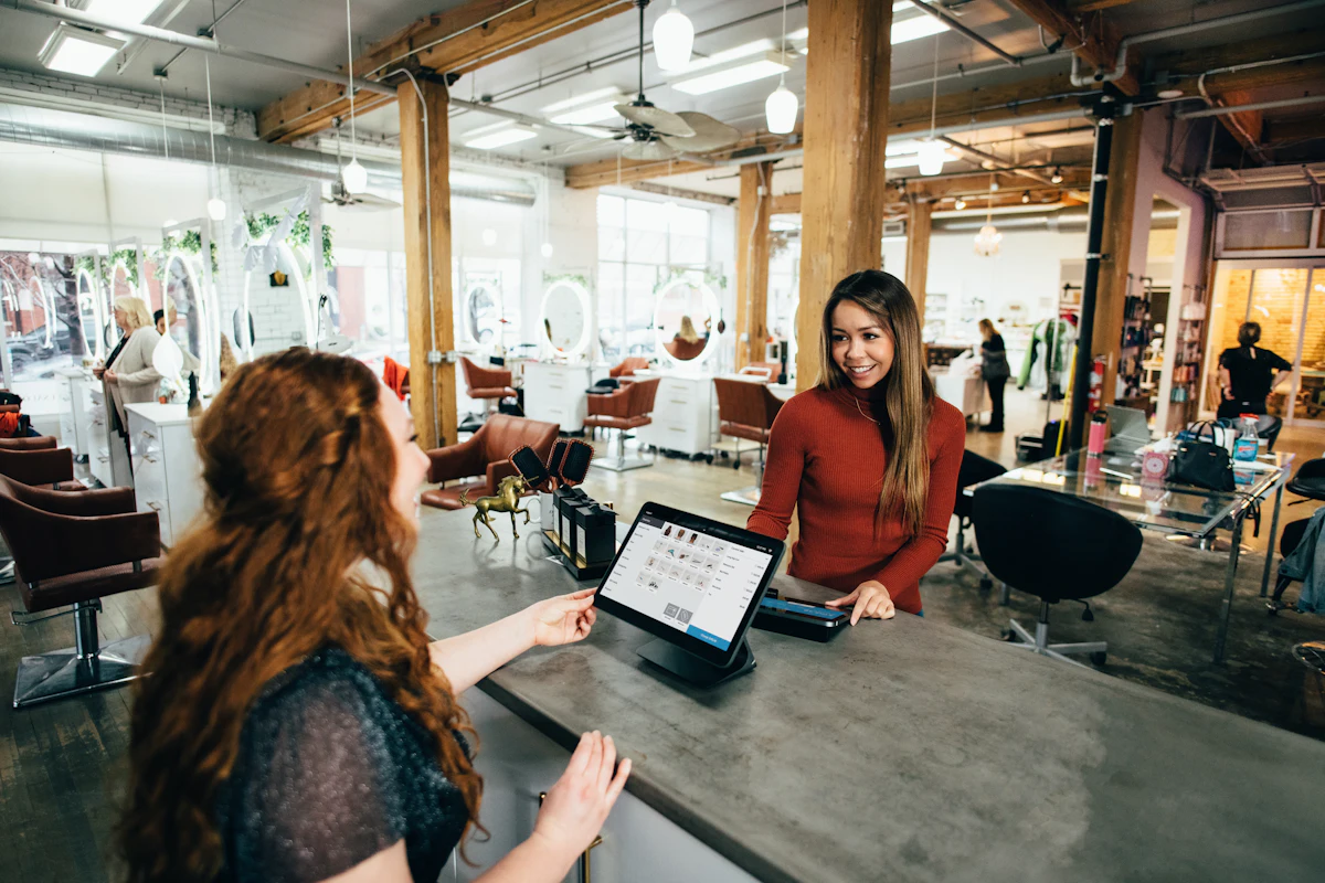 Customer chatting with a smiling receptionist at a salon front desk with a tablet register