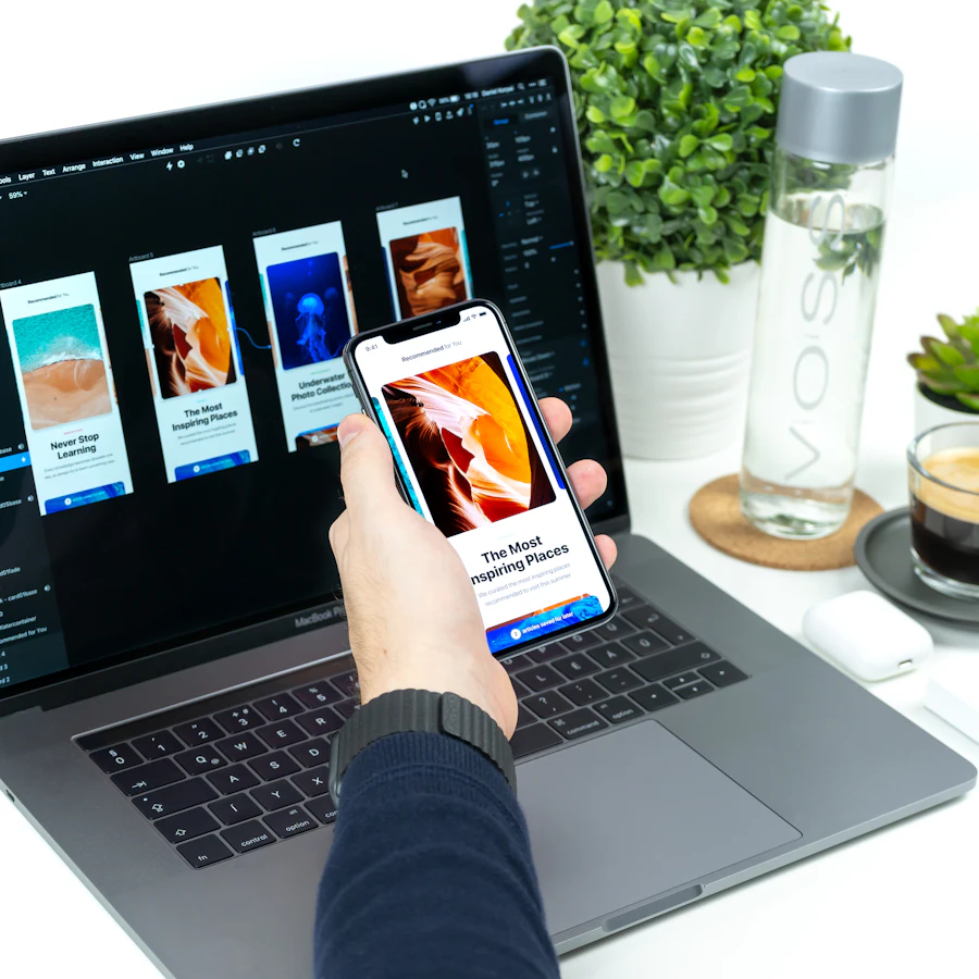 Hand holding a smartphone in front of a laptop that shows the same app's screens laid out in a design file